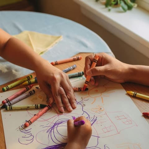 Close-up from above of a young child's hands and a parent's hands drawing with crayons on paper